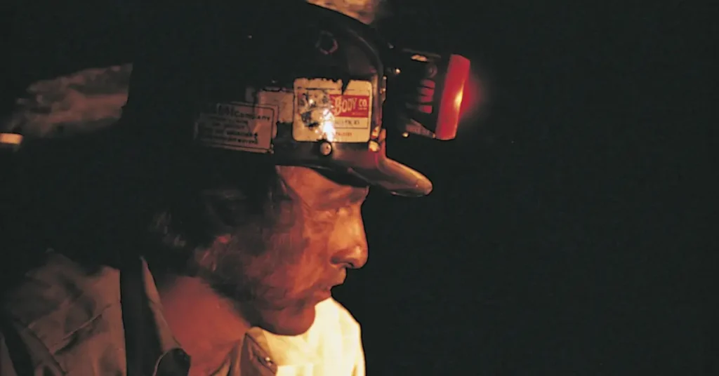 A coal miner wearing a hard hat with a headlamp works in a dark underground mine. The dim lighting highlights the miner’s focused expression and rugged environment, emphasizing the physically demanding and hazardous nature of the job.