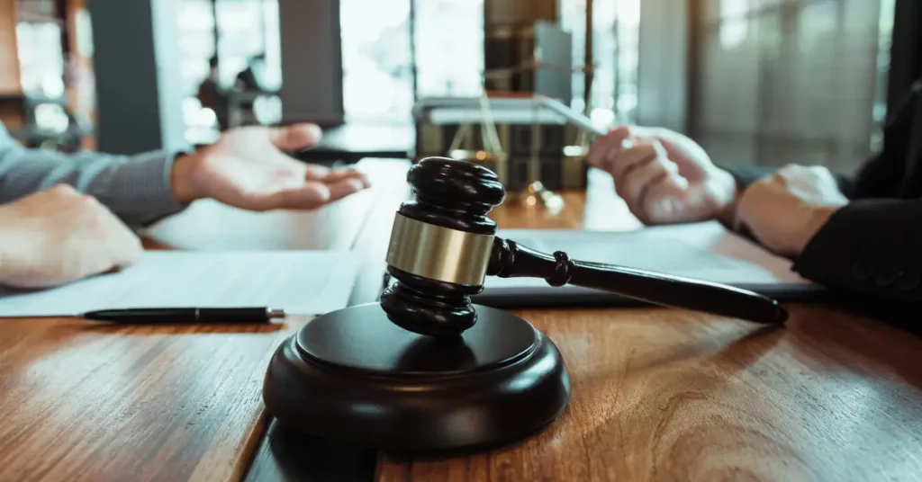 A close-up of a judge’s gavel on a wooden table, with two individuals in discussion blurred in the background. Legal documents, a pen, and the scales of justice are also visible, indicating a formal legal meeting or court proceeding.