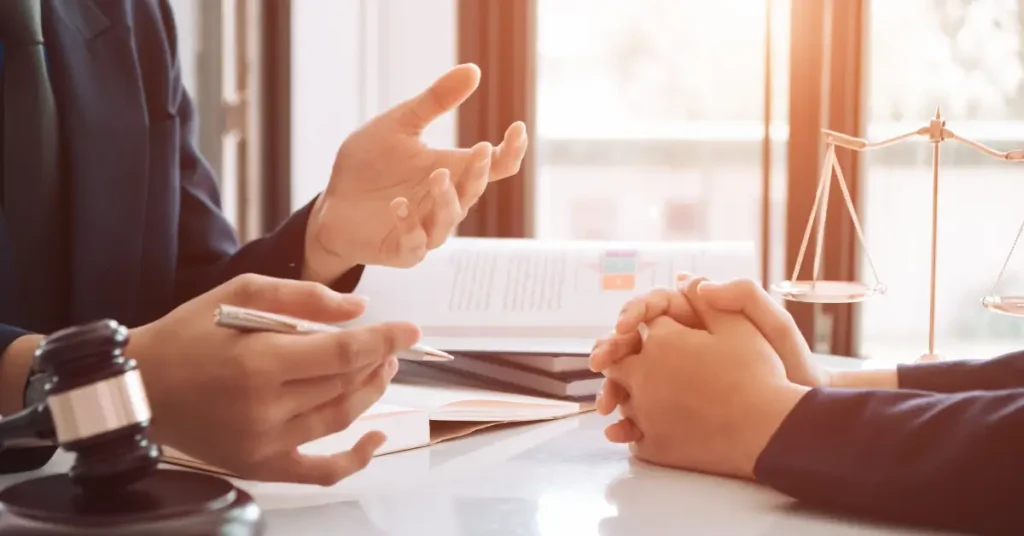 Two people in a professional setting are seated across from each other at a desk, engaged in a serious conversation; one is gesturing while the other listens with hands clasped. A gavel and scales of justice are visible, indicating a legal or consultation environment.