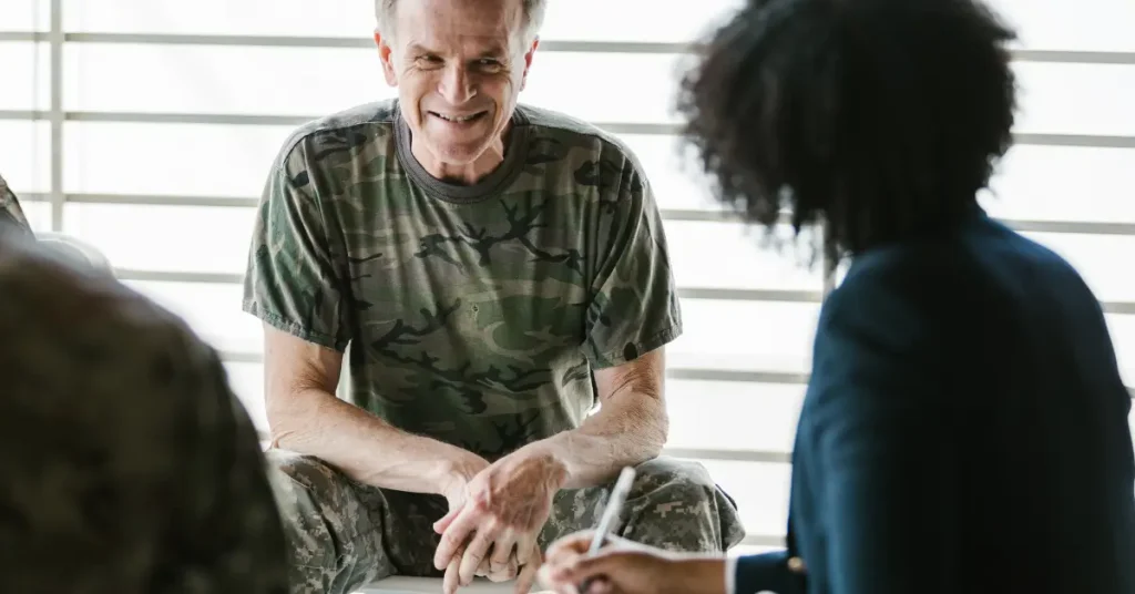 A smiling older man in military camouflage sits in a brightly lit room, engaged in conversation with a woman taking notes. The scene suggests a supportive setting, possibly a therapy or counseling session for veterans.