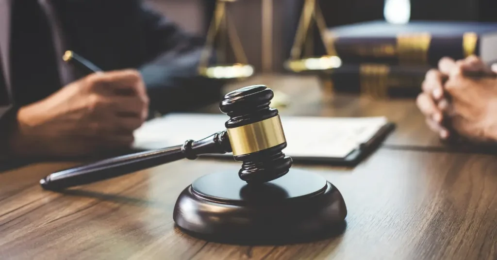 A judge’s gavel rests on a wooden desk with legal documents, scales of justice, and two people sitting across from each other in the background. The setting suggests a courtroom or legal consultation, symbolizing justice, legal proceedings, or a court decision.
