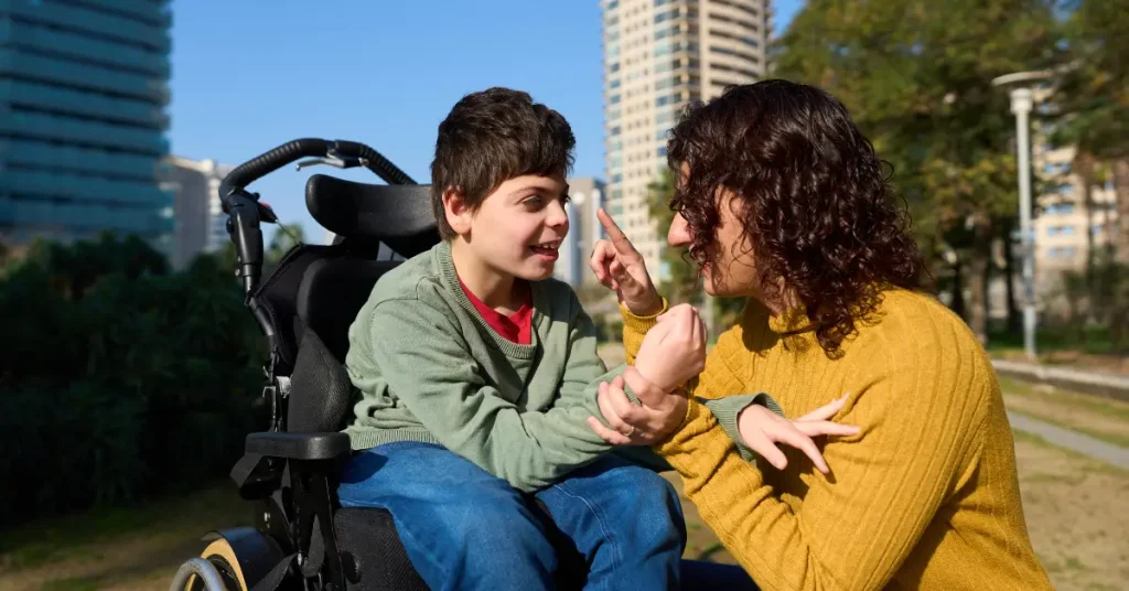 A young boy in a wheelchair interacts joyfully with a woman, possibly his mother or caregiver, in an outdoor park setting. Both are smiling and using expressive hand gestures, suggesting a strong emotional connection and communication. Tall buildings and trees are visible in the background.