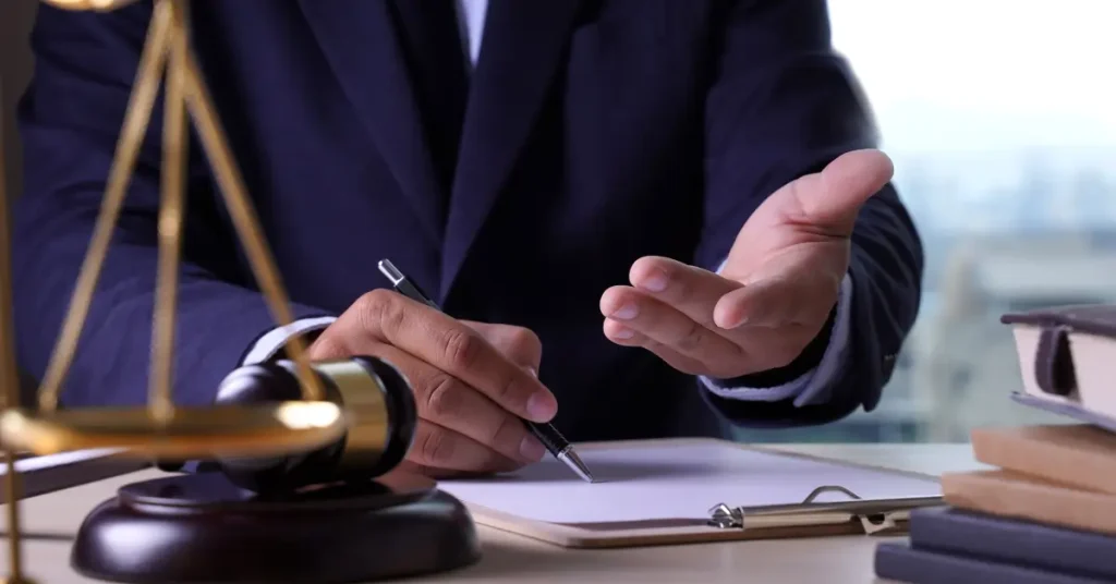 A person in a dark suit sits at a desk with a clipboard, gesturing with one hand while holding a pen in the other. A gavel, legal scales, and stacked books are visible on the desk, suggesting a legal or courtroom setting, possibly a discussion or consultation with a lawyer or judge.