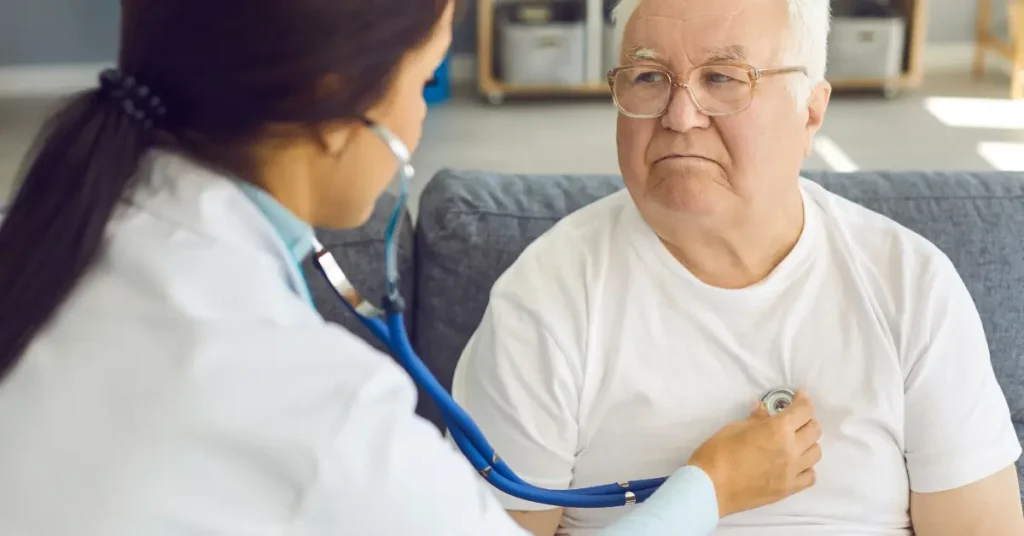 A female doctor uses a stethoscope to listen to the chest of an elderly man wearing glasses and a white T-shirt. They are seated in a well-lit room, suggesting a medical check-up or evaluation, possibly related to heart health.