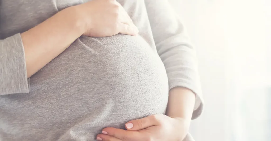 A close-up of a pregnant woman wearing a gray shirt gently cradling her belly with both hands. The soft lighting and serene setting emphasize themes of motherhood, pregnancy, and maternal care.
